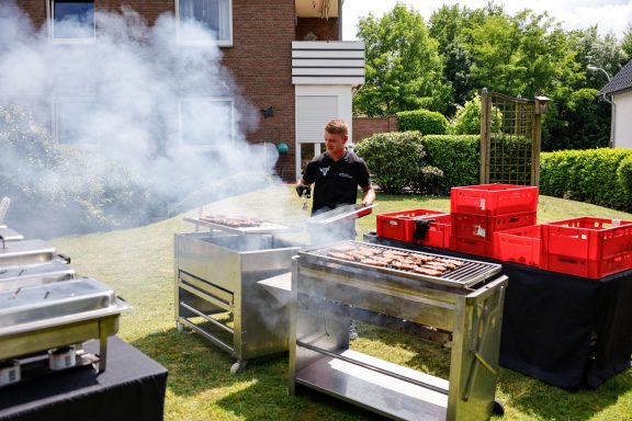 Grillmeister bereitet Fleisch auf einem Grill im Freien vor, umgeben von grüner Landschaft.