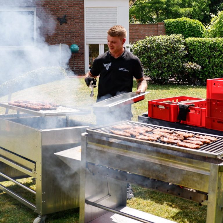 Ein Mann grillt Würstchen auf einem Grill im Freien, umgeben von Rasen und Bäumen.