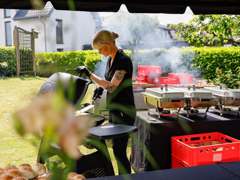Frau grillt im Garten bei einer Veranstaltung, Rauch von den Grillgeräten aufsteigend.